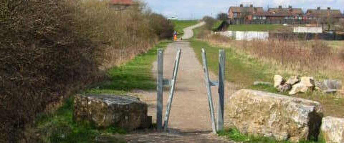 A rural pathway leading to a gated entrance with a stone wall on one side and a residential area in the background.