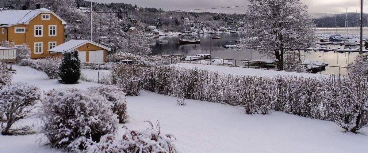 A snow-covered landscape with a yellow house, a frozen lake, and boats docked.