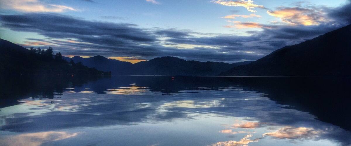 A calm lake at dusk with mountains in the background and a cloudy sky.
