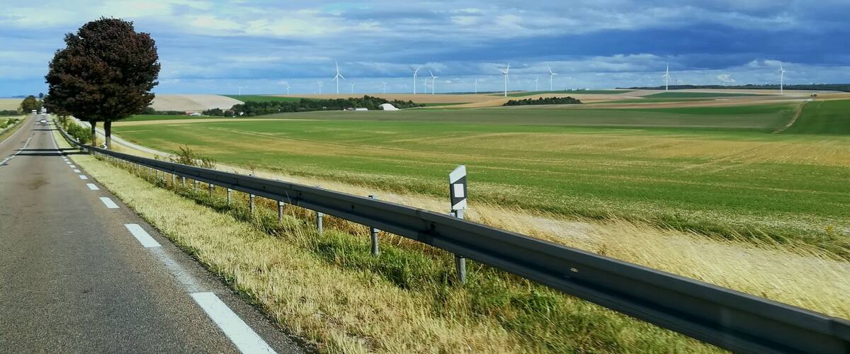 A rural road with a tree on the left, wind turbines in the distance, and a clear sky.