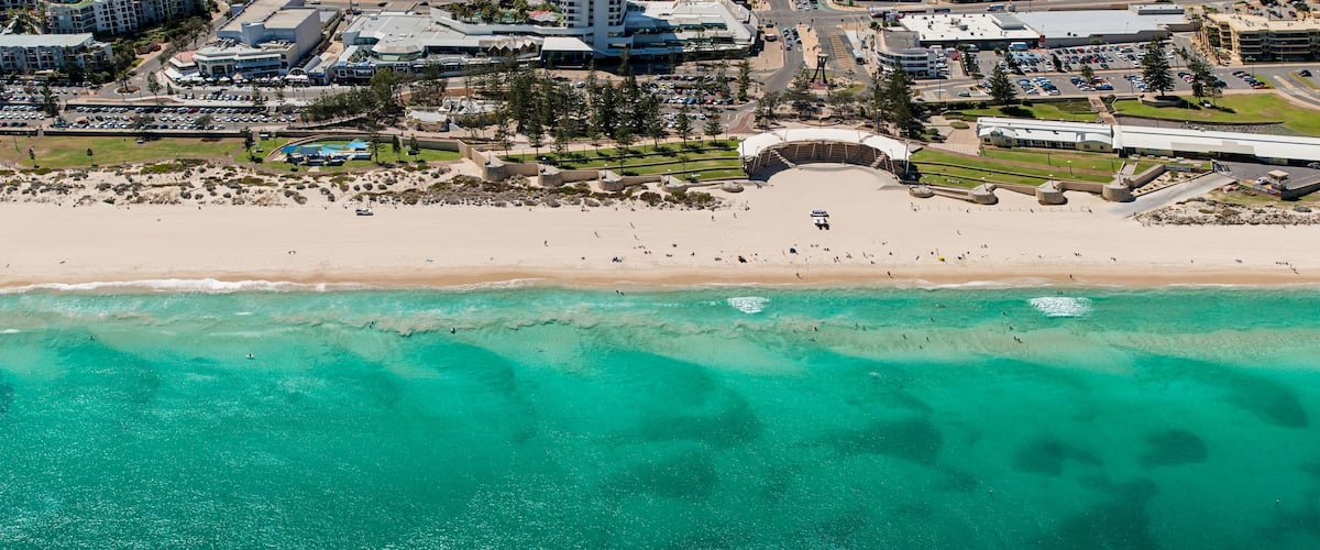 An aerial view of a beach with clear turquoise waters, a sandy shore, and a large building complex in the background.