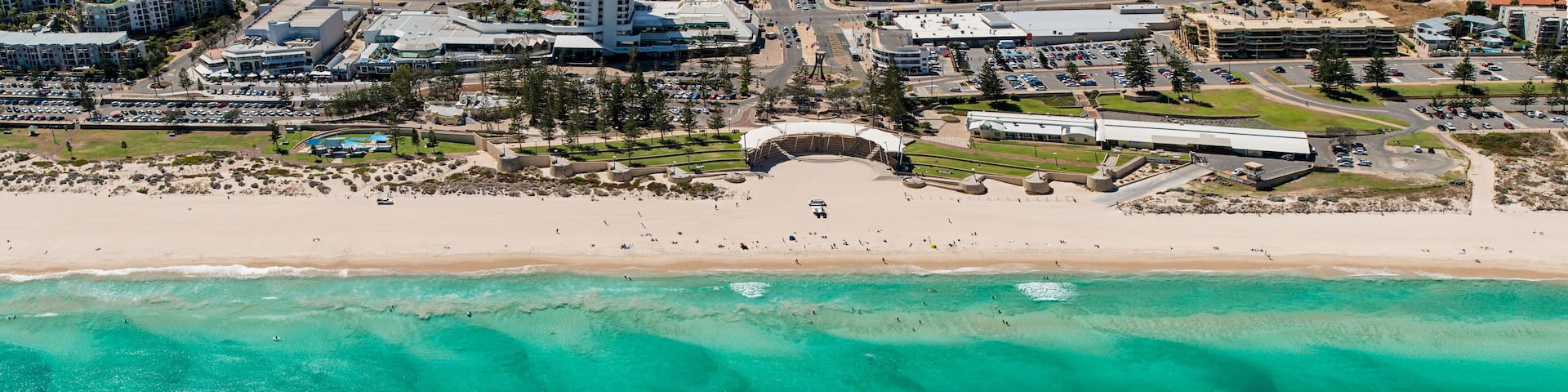An aerial view of a beach with clear turquoise waters, a sandy shore, and a large building complex in the background.