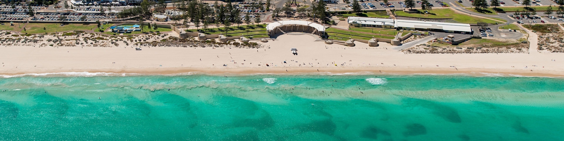 Vista aérea de una playa con aguas turquesas cristalinas, una orilla arenosa y un complejo arquitectónico grande al fondo.