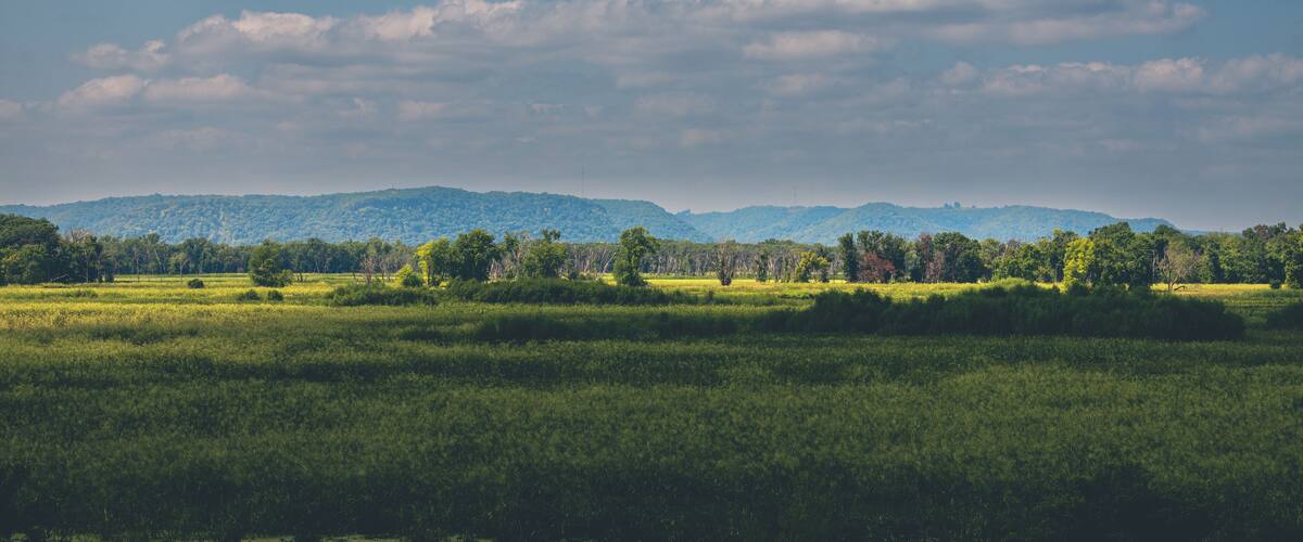 A lush green landscape with a river in the foreground, rolling hills in the background, and a partly cloudy sky.