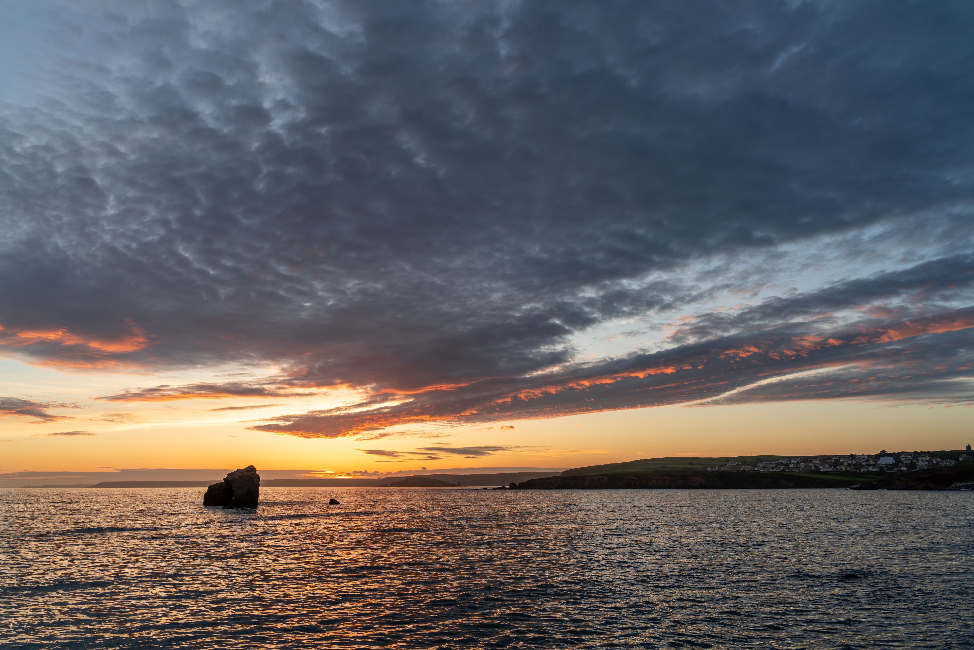 Un coucher de soleil sur une étendue d'eau, avec une formation rocheuse au premier plan et une côte lointaine parsemée de bâtiments.