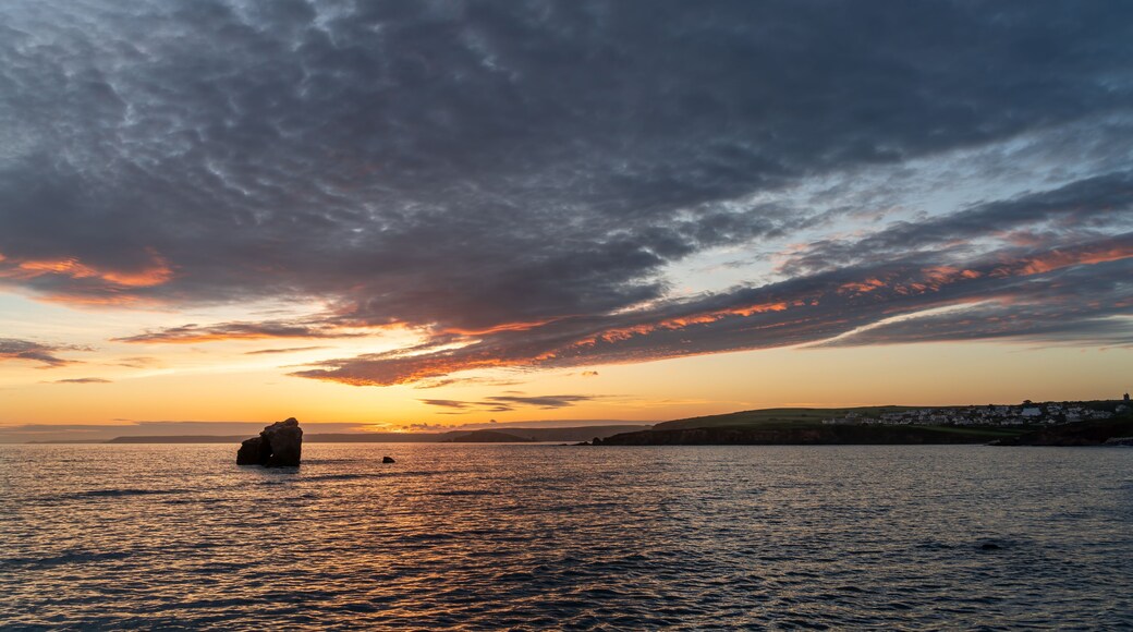 Un coucher de soleil sur une étendue d'eau, avec une formation rocheuse au premier plan et une côte lointaine parsemée de bâtiments.