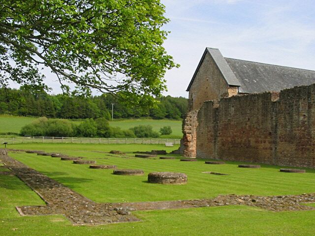 A historic stone building with a thatched roof, surrounded by a grassy field with ancient stone structures.