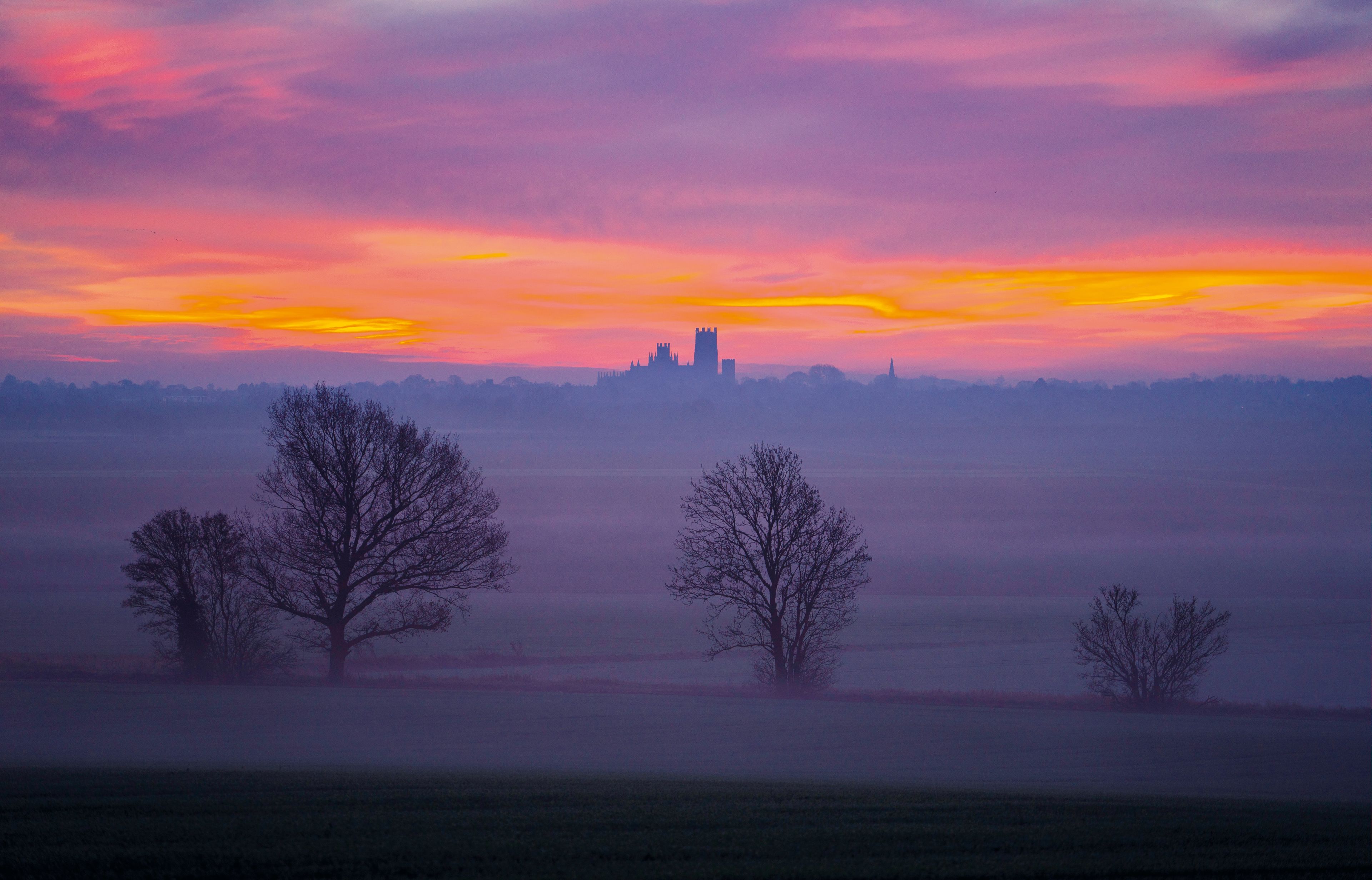 Paisagem com silhueta de castelo contra um céu de pôr do sol colorido, rodeado por árvores secas e um campo nevoento.