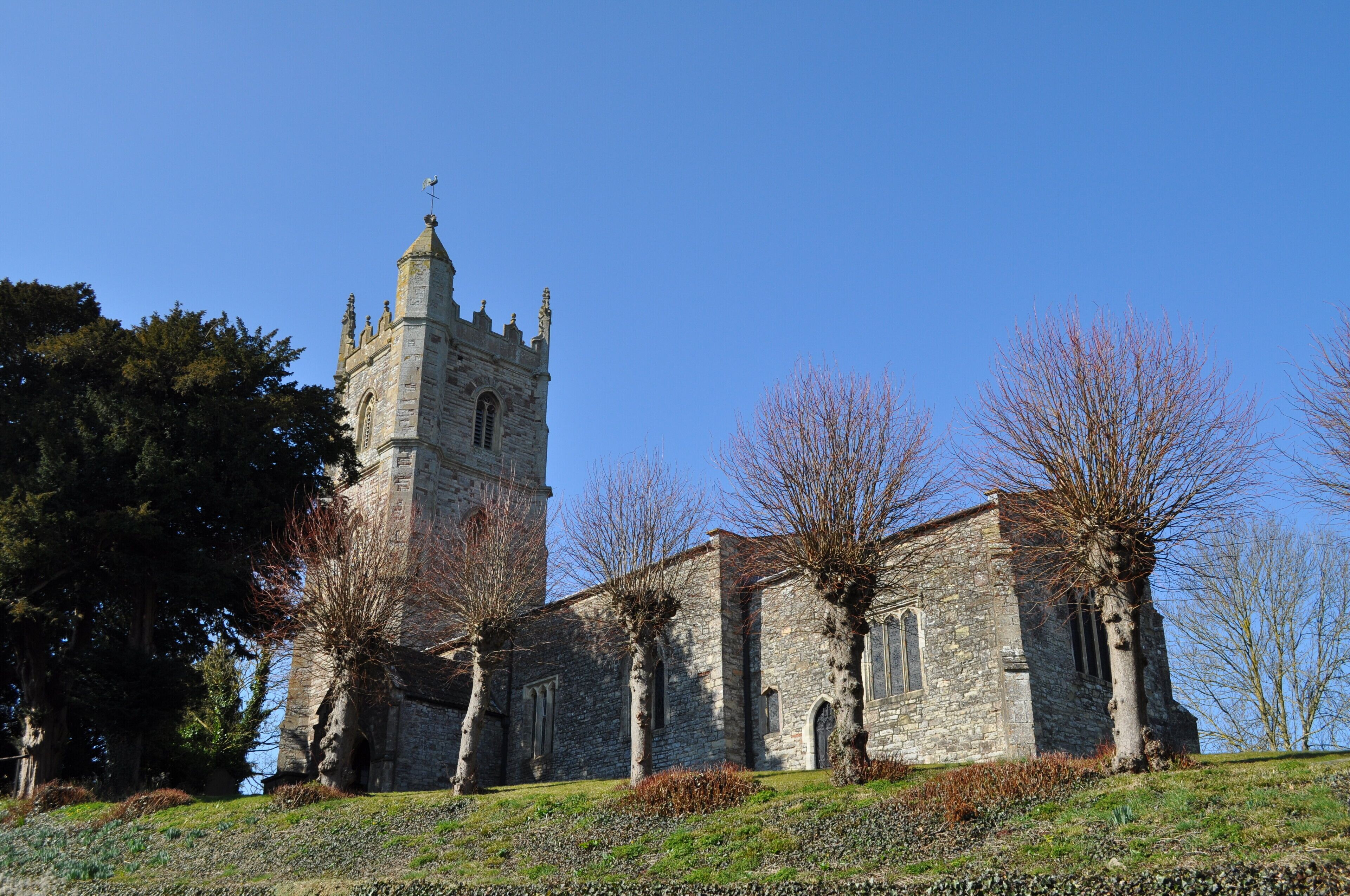 A historic stone church with a tall bell tower and leafless trees in a sunny landscape.