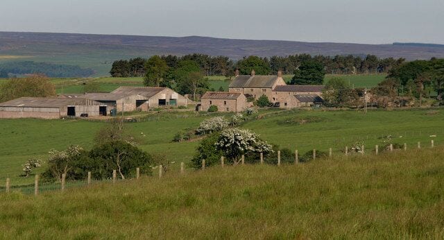 A rural landscape with farm buildings, green fields, and distant hills.