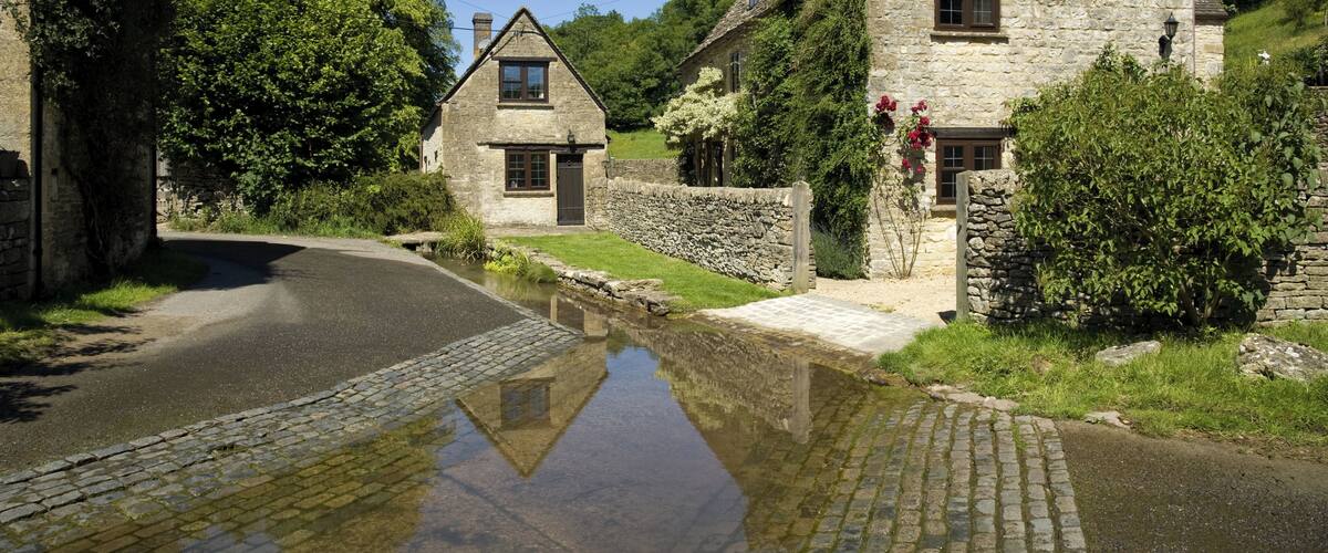 A stone cottage with a pond in front, surrounded by greenery and trees.