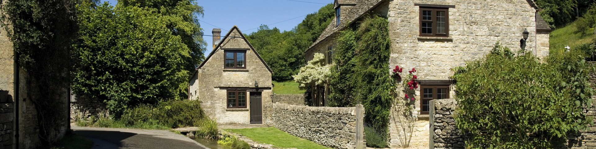 A stone cottage with a pond in front, surrounded by greenery and trees.