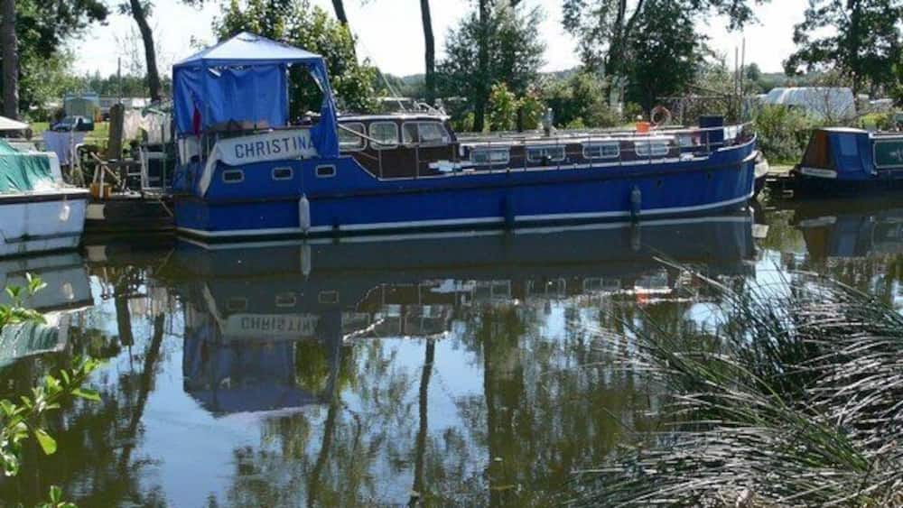 A blue boat named CHRISTINA docked at a calm water body surrounded by trees and greenery.