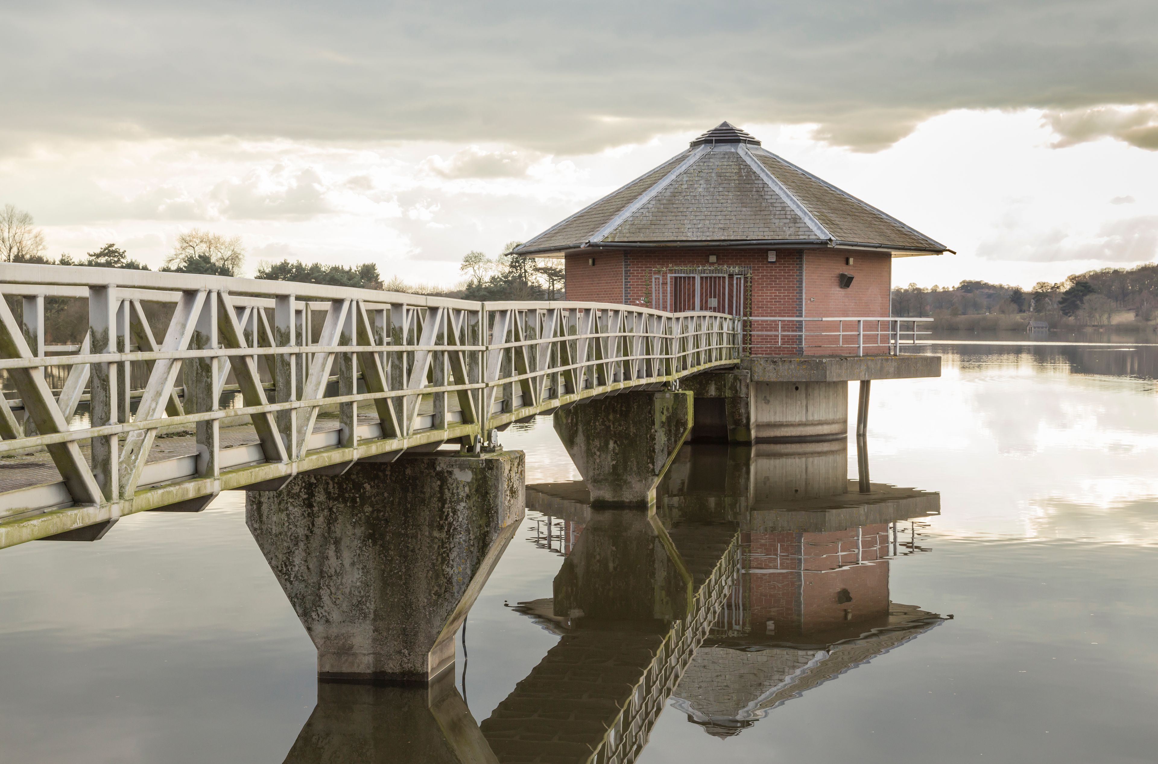 A bridge over a calm lake with a small brick building on stilts in the background.