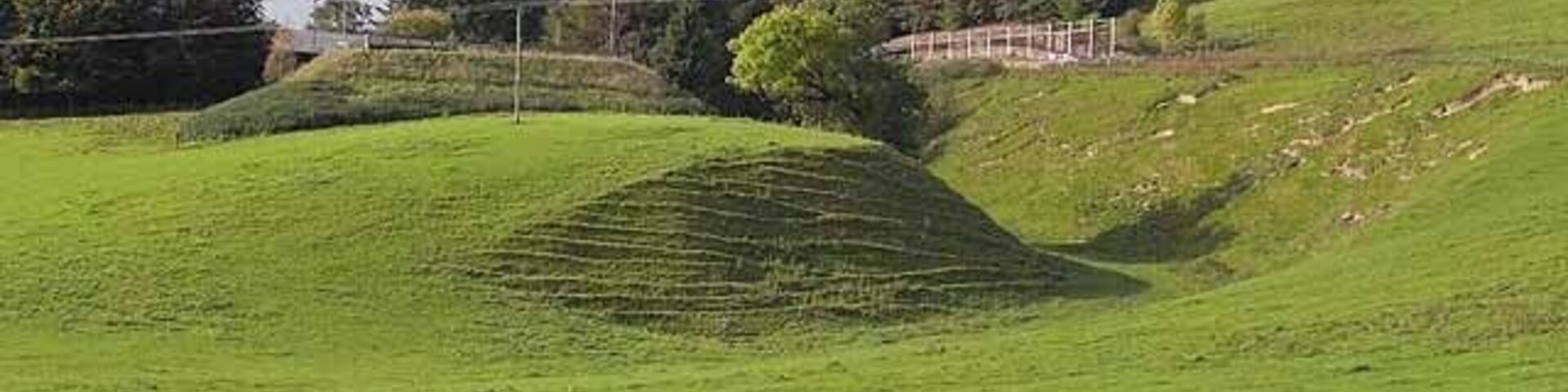 Un campo verde con una colina curva y una valla al fondo.