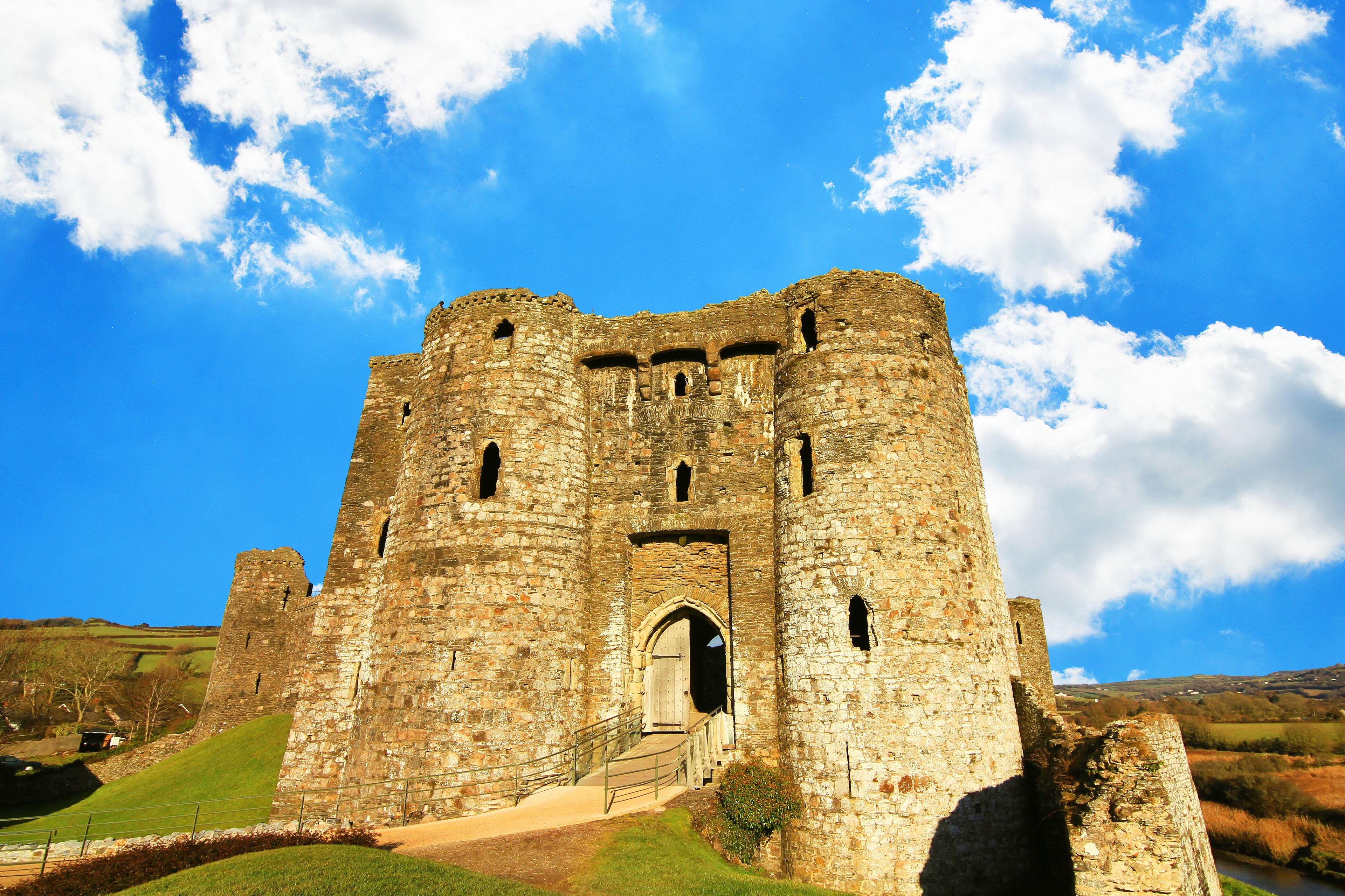 A historic stone castle with multiple towers and a central entrance, surrounded by a grassy landscape under a clear sky.