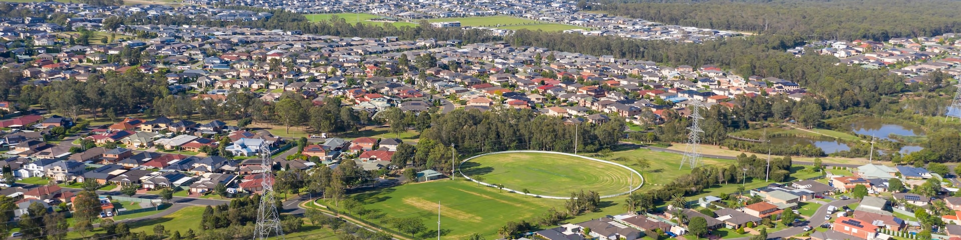 Uma área residencial com casas, ruas, um amplo campo verde e colinas ao longe.