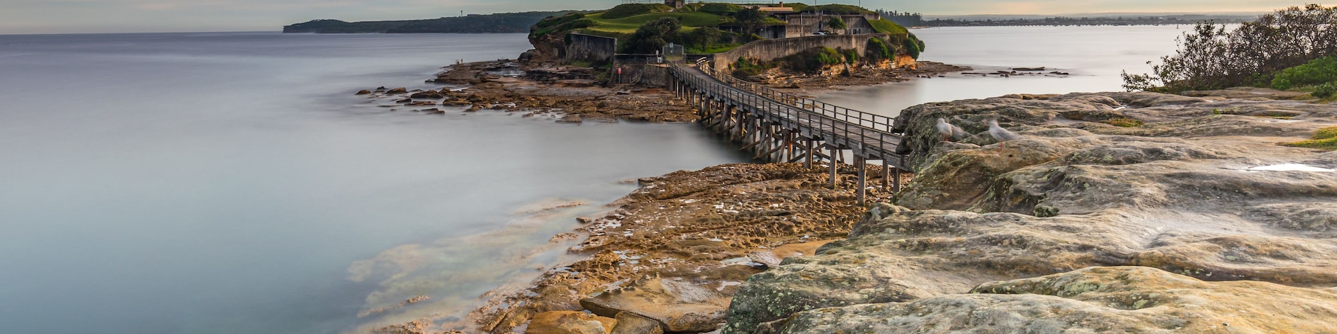 Paisagem costeira com falésias rochosas, um cais e uma ilha distante com uma fortificação.