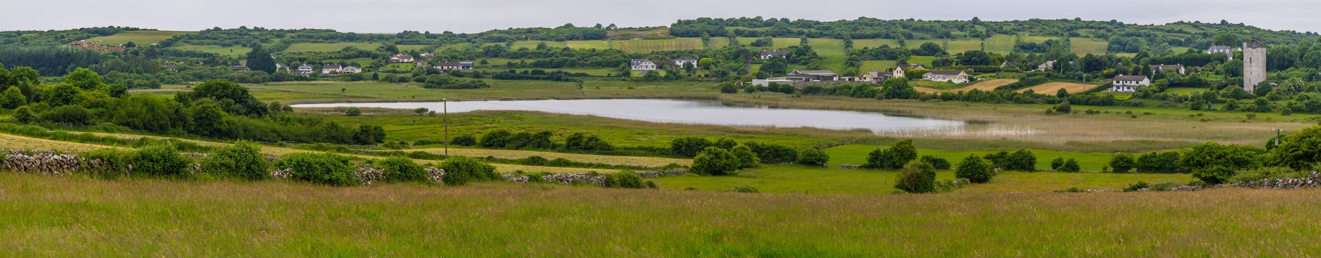 Un paysage rural avec un lac, des collines ondoyantes, des maisons dispersées et un imposant édifice en forme de tour.