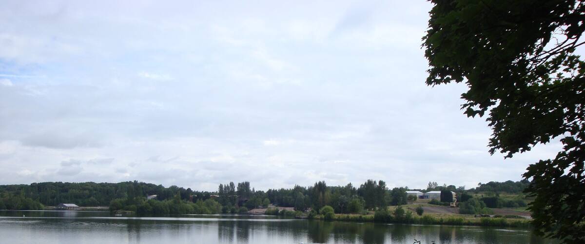 A calm lake with a forested shoreline, a small building, and a fence in the foreground.