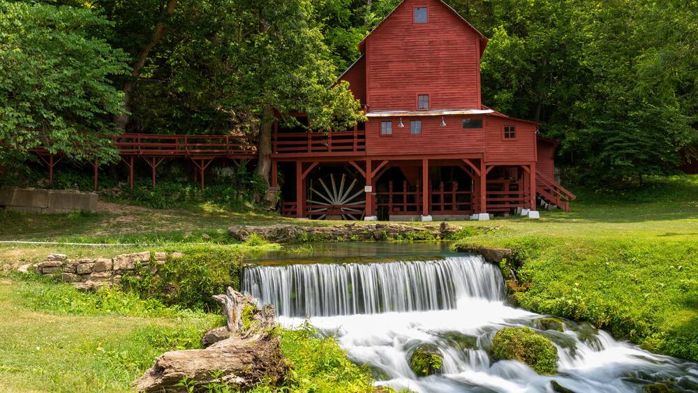 A red watermill with a wooden wheel, surrounded by lush greenery and a small waterfall.