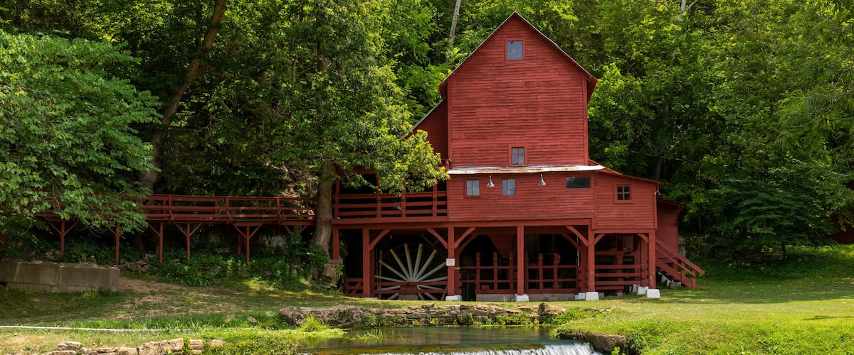 A red watermill with a wooden wheel, surrounded by lush greenery and a small waterfall.