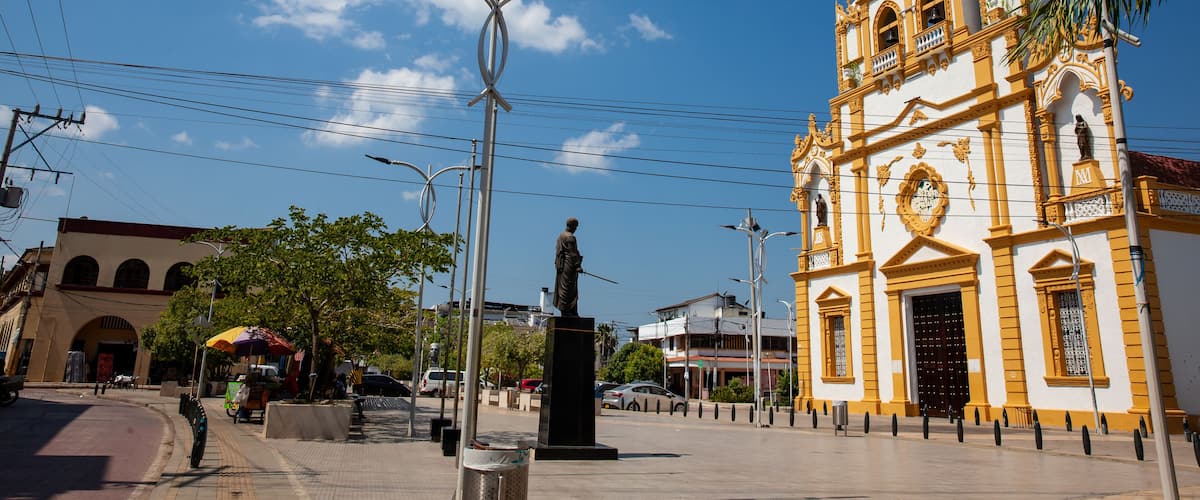 A historic church with a statue in front, surrounded by a plaza with palm trees and street lamps.