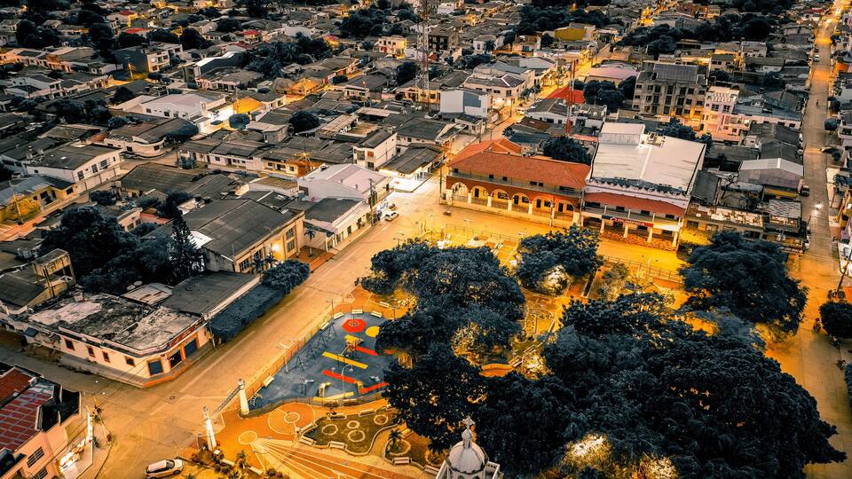 An aerial view of a cityscape with a prominent building with a red roof, surrounded by trees and a sports court.