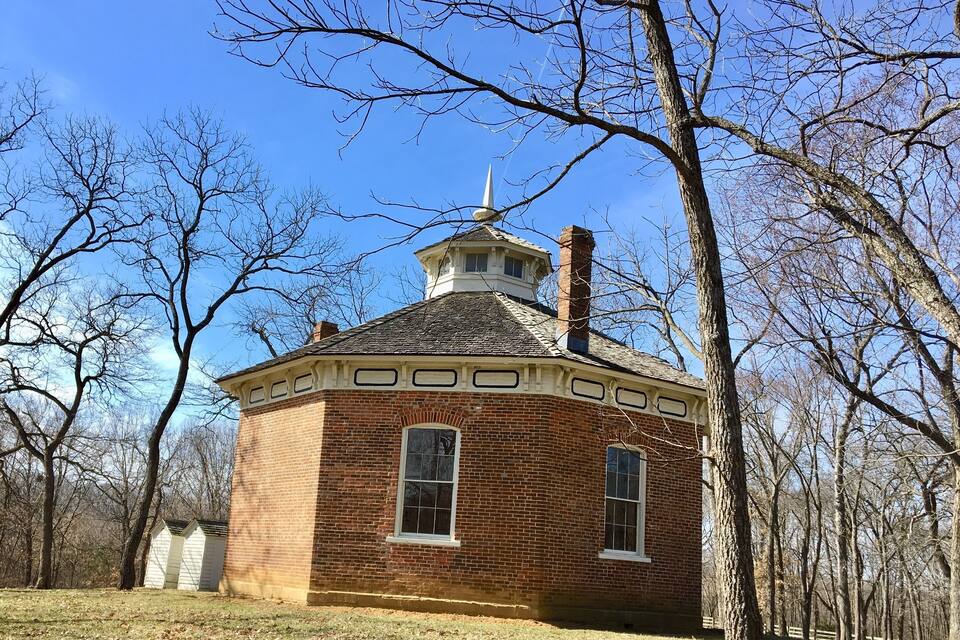 A brick building with a tower and a small shed in a wooded area.