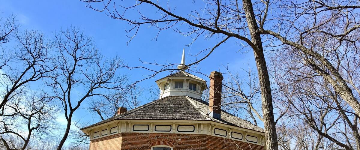 A brick building with a tower and a small shed in a wooded area.