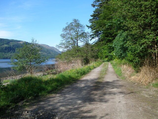 A gravel path beside a lake, surrounded by trees and hills.