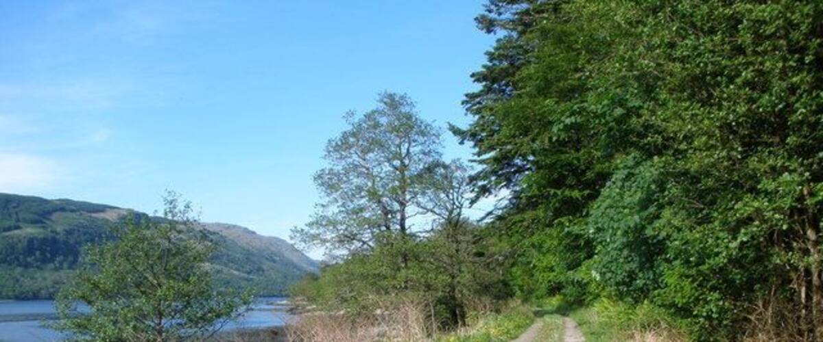 A gravel path beside a lake, surrounded by trees and hills.