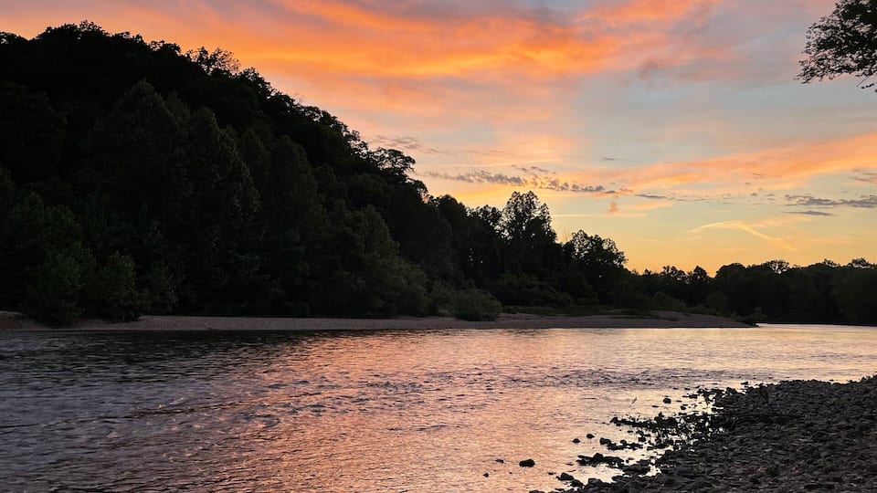 A riverbank with a rocky shoreline and a forested hill in the background.