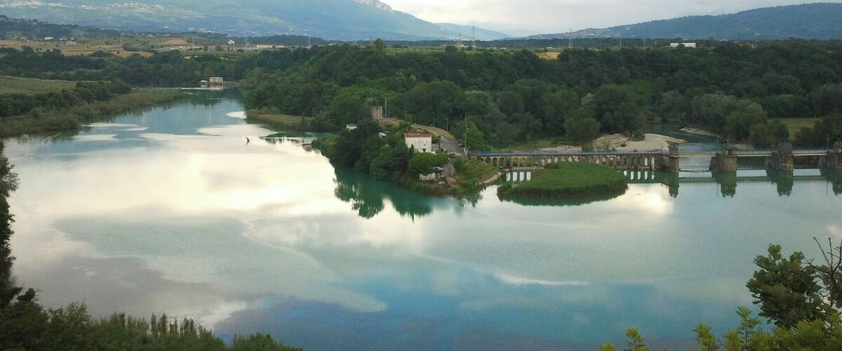 A calm river with a bridge, surrounded by lush greenery and mountains in the background.