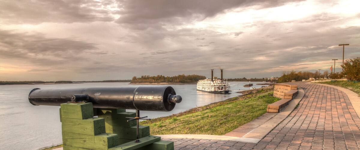 A cannon on a brick platform by a river, with a steamboat in the background.