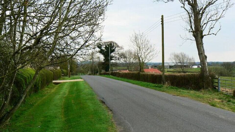 A rural road flanked by hedgerows and trees, leading towards a distant red-roofed building.