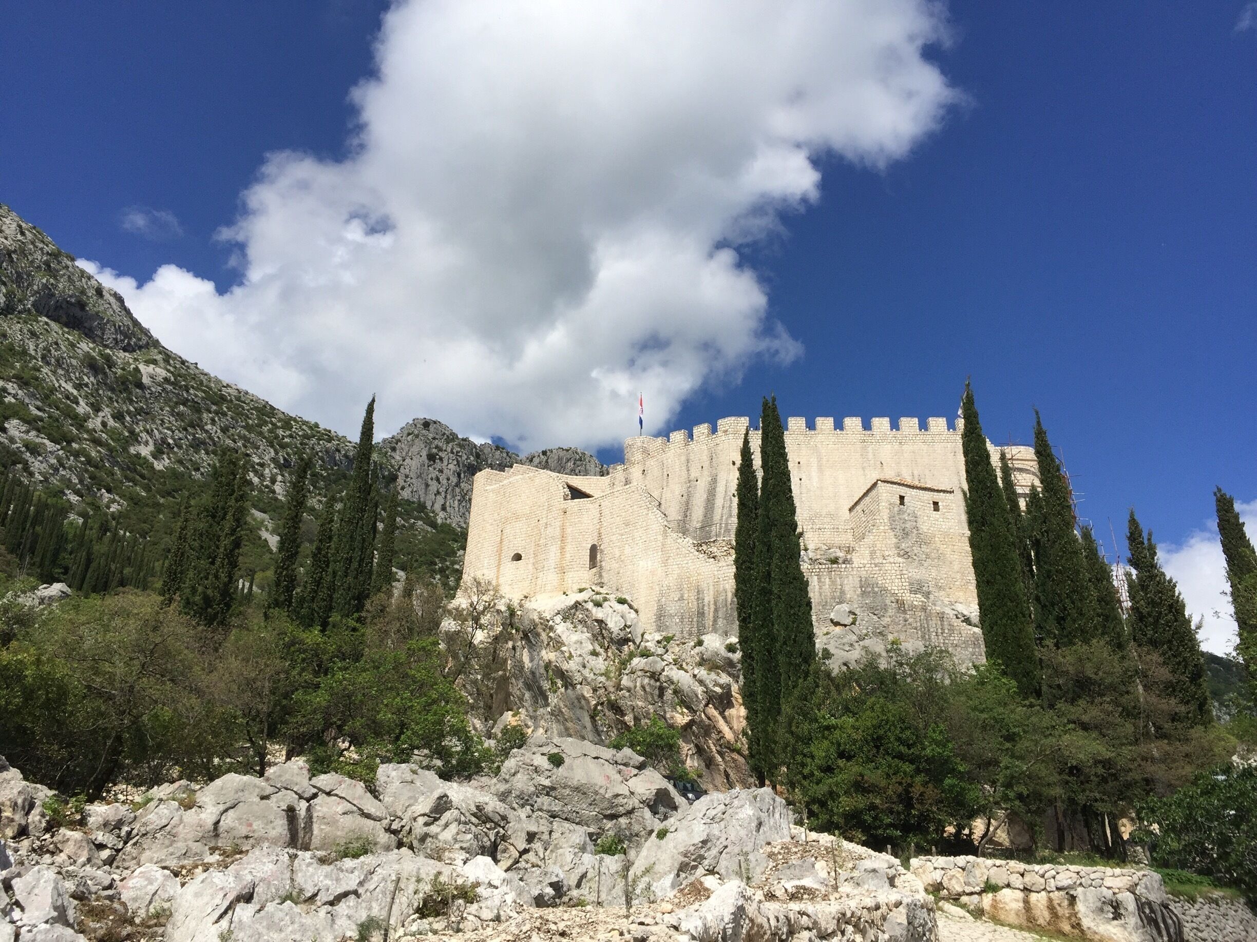 A historic stone fortress with battlements, surrounded by rocky terrain and cypress trees.