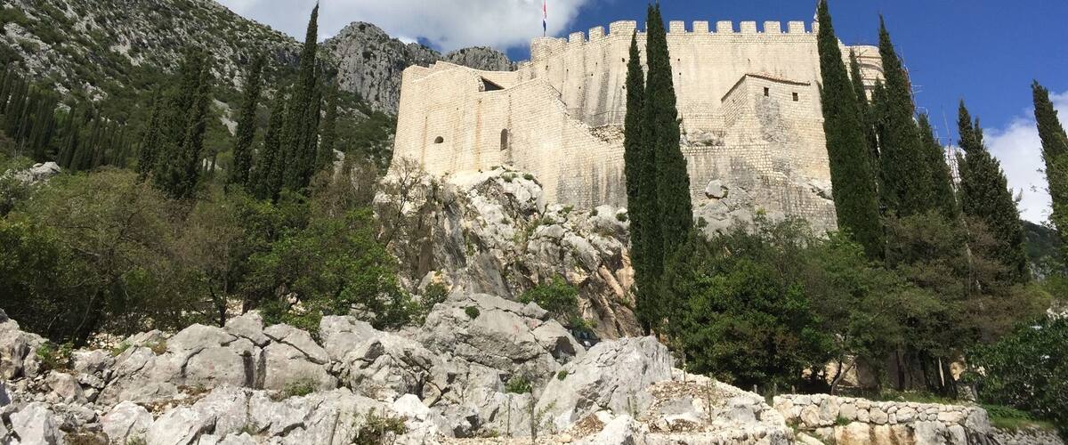 A historic stone fortress with battlements, surrounded by rocky terrain and cypress trees.