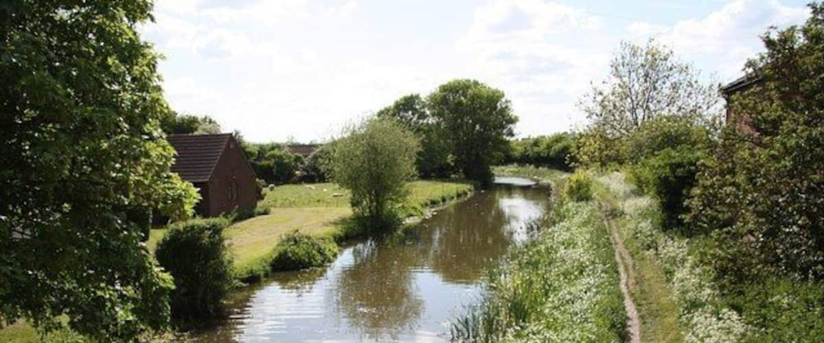 A narrow river with a wooden walkway alongside, surrounded by greenery and a small building in the distance.