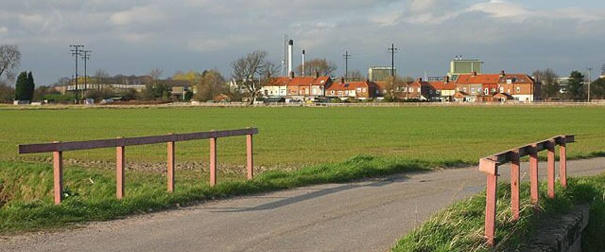 A rural road with wooden guardrails, a green field, and buildings in the background.
