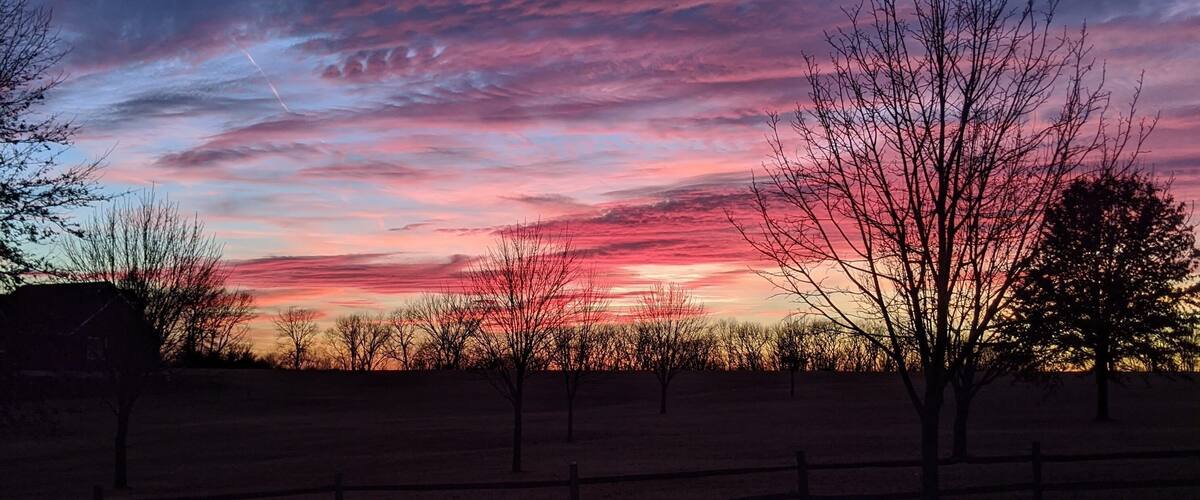 A sunset over a rural landscape with bare trees and a wooden fence.