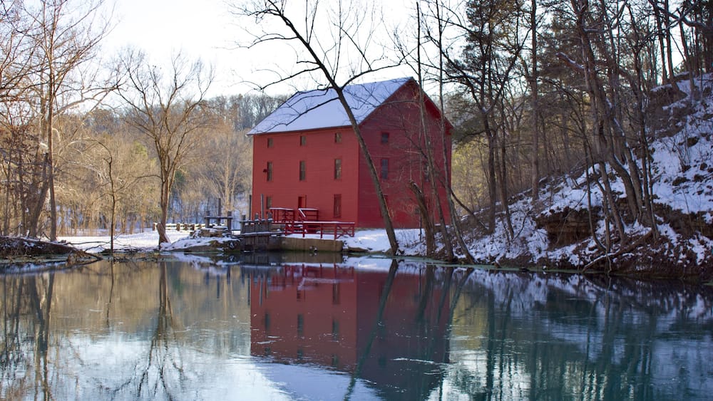 A red barn by a calm lake with snow on the ground and trees in the background.
