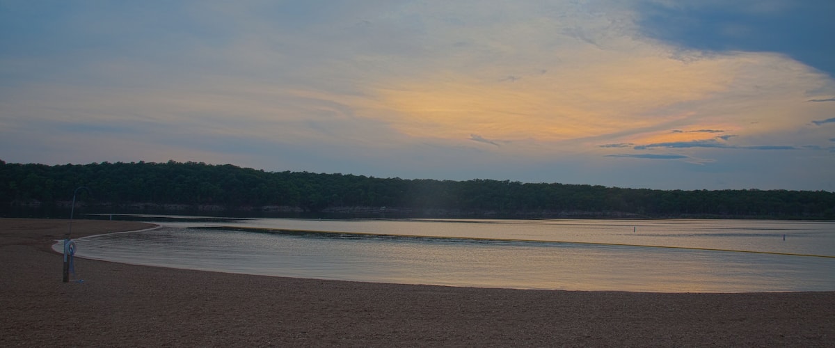 A beach with a curved shoreline, a sandy foreground, and a forested background under a partly cloudy sky.