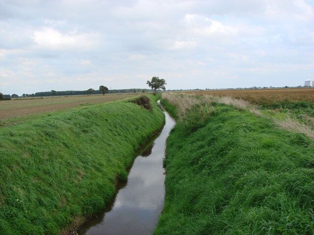 A narrow water channel flanked by lush green grass fields under a cloudy sky.