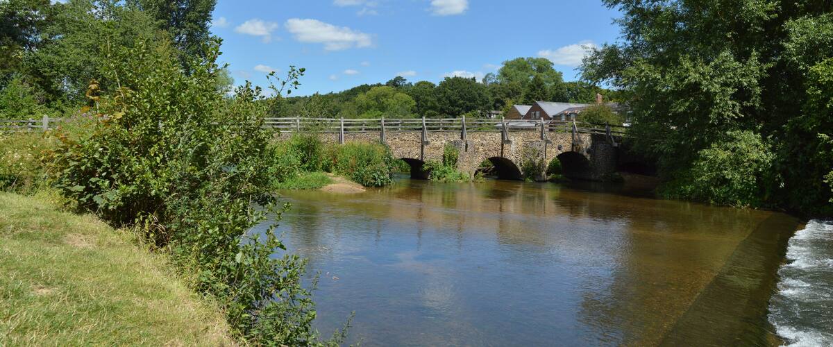 A stone bridge over a calm river, surrounded by lush greenery and a clear blue sky.