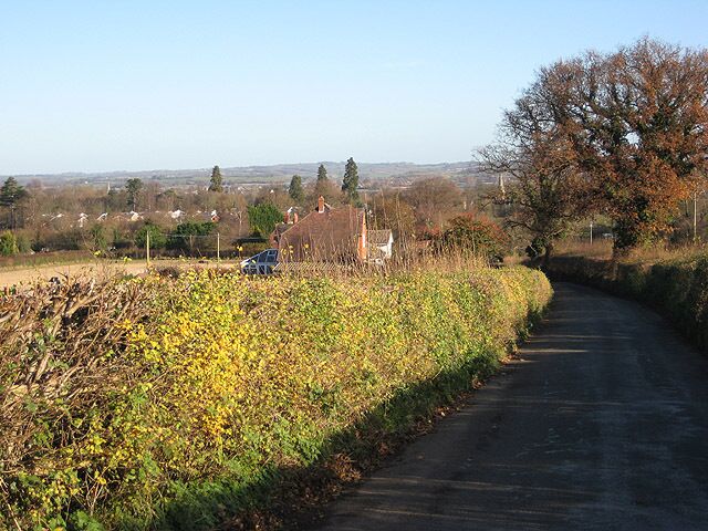 A narrow rural road with a car parked on the side, surrounded by fields and trees.