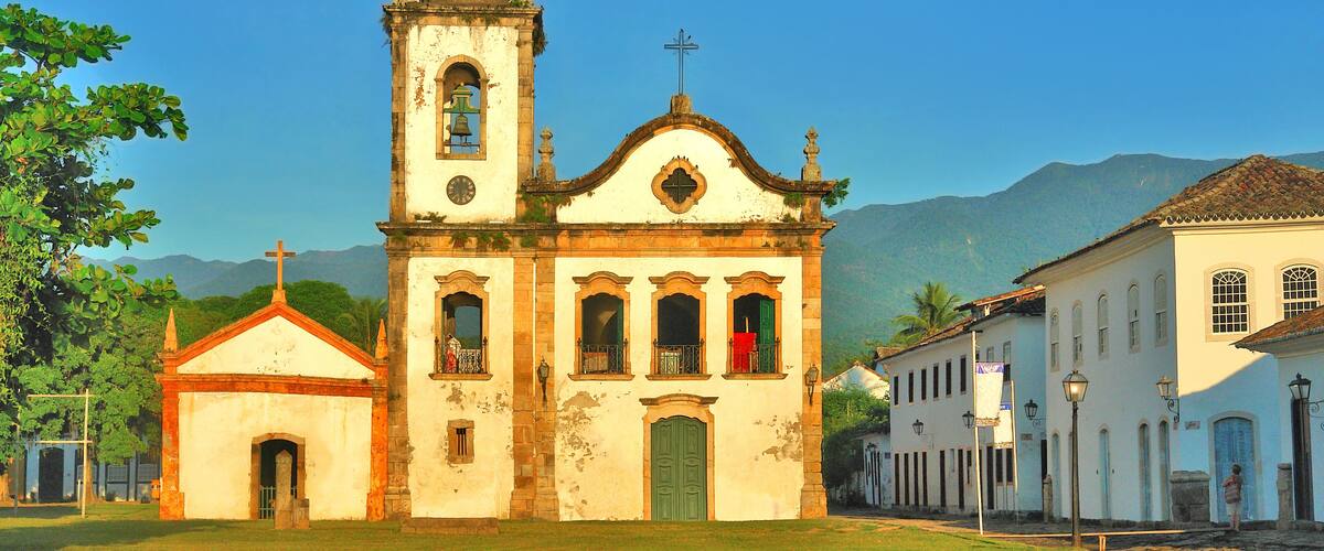 A historic church with a bell tower, surrounded by lush greenery and mountains in the background.