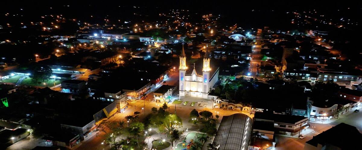 An aerial view of a city at night with illuminated buildings, a prominent church with two spires, and a well-lit square with trees and a fountain.