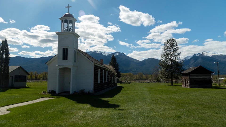 A white church with a bell tower and a cross, surrounded by a grassy field and mountains in the background.