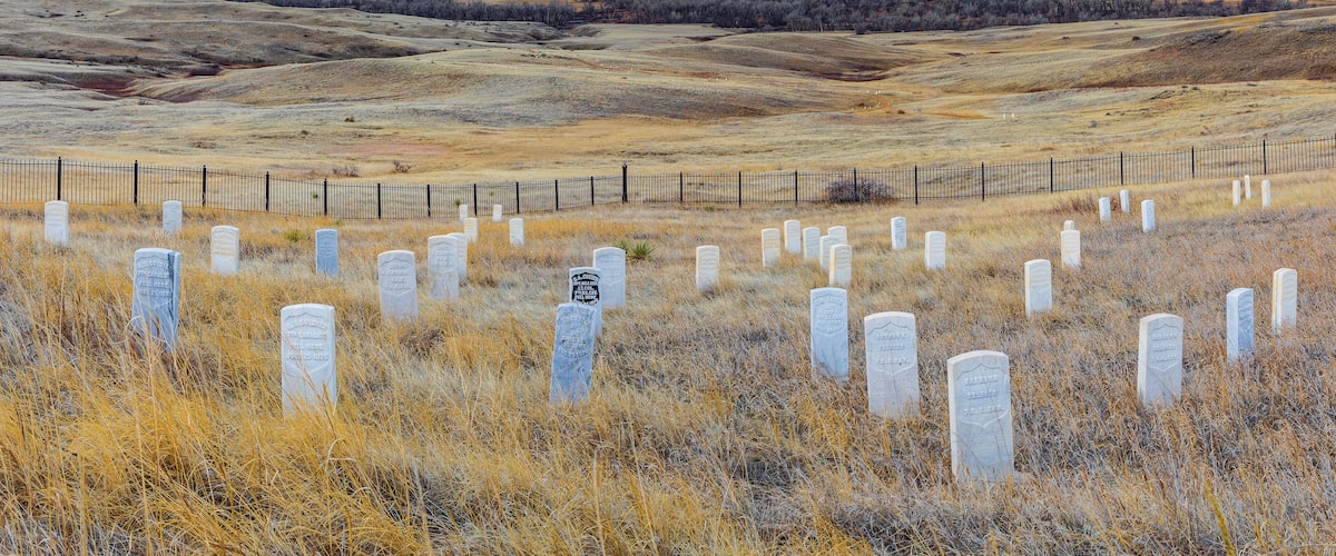 A cemetery with white headstones in a dry grass field, surrounded by a fence.